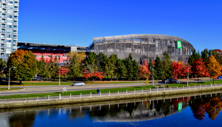 TD Place stadium seen across the Rideau Canal, with trees in autumn colour.