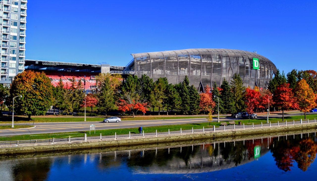 TD Place stadium seen across the Rideau Canal, with trees in autumn colour.