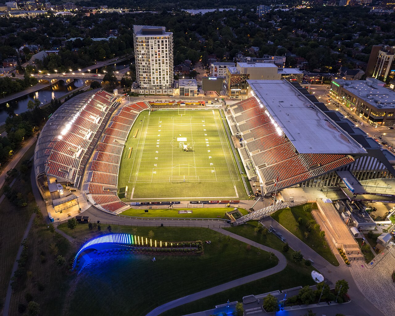 Aerial view of TD Place stadium lit up at night, with the field and stands visible.
