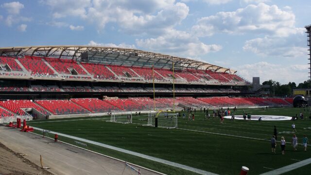 The stadium at TD Place in Ottawa: rows of red seats under a curved roof, with goalposts and people on the green field on a sunny day